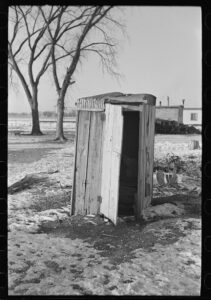 Black and white photo of an outhouse with snow on the ground and trees in the background.