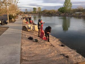 A line of people fishing at an artificial pond with a concrete sidewalk behind them.