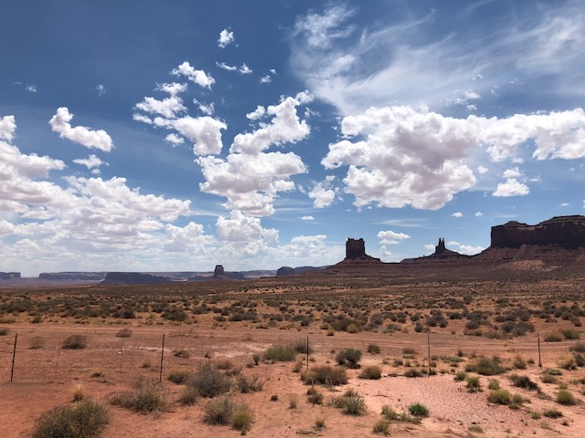 A panoramic view of Monument Valley, featuring red sandstone buttes and mesas rising from the desert floor under a bright blue sky with fluffy white clouds. The rugged landscape is sparsely vegetated with shrubs and grasses.