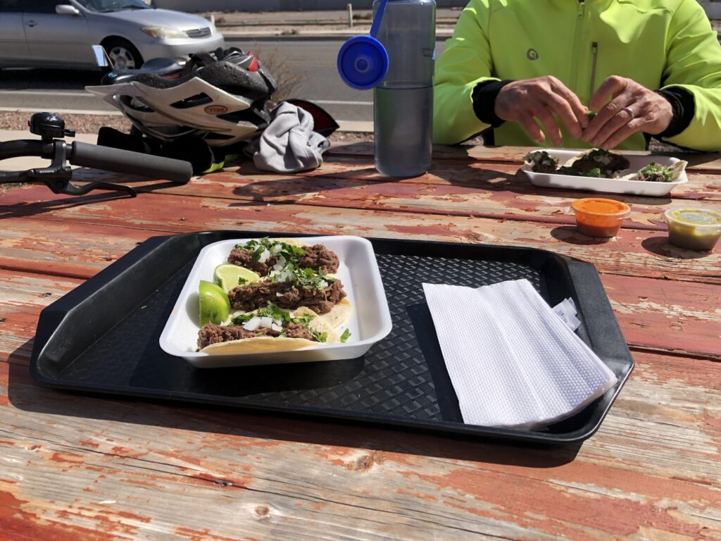 A plate of tacos on a black tray sits on a weathered wooden table. The tacos are garnished with cilantro and lime wedges. In the background, a person in a bright green jacket is eating from another plate. A bicycle helmet, water bottle, and car are visible, suggesting an outdoor dining setting.