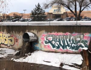 A concrete drainage channel or culvert with graffiti art on its walls, featuring colorful tags in pink and gray tones. Above the culvert is a parking lot with a Ross store visible in the background. The scene is partially covered in snow