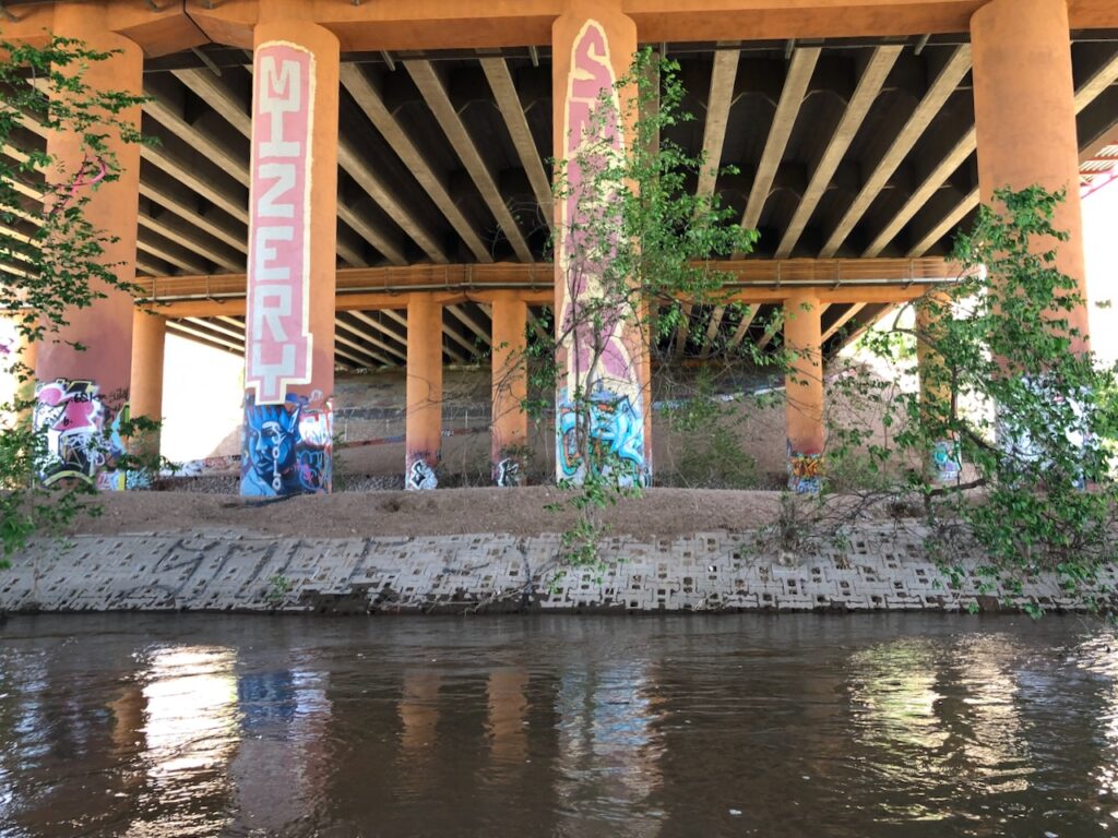 The underside of a bridge, with orange columns covered with graffit and a river in the foreground.