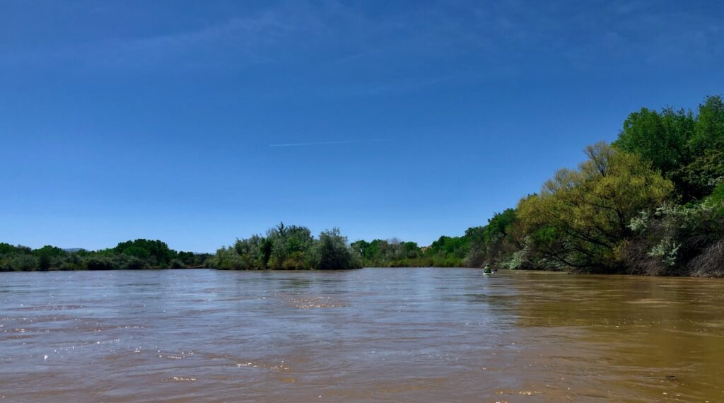 Brown river, blue sky, trees flanking the river, a paddler paddling a boat in the distance