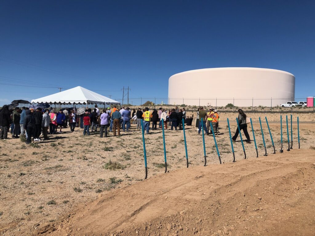 Large tan water tank reservoir in the background, party tent in mid-ground, people milling about, a line of shovels in a pile of dirt in the foreground.