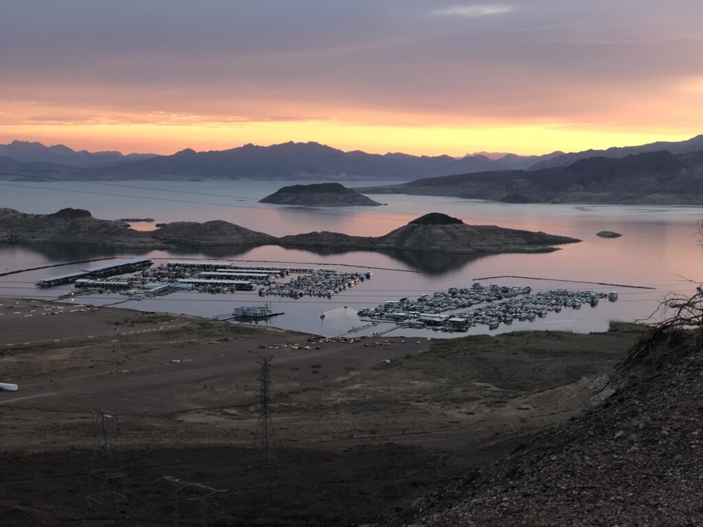 Scenic sunrise over a large lake with mountains in the background. In the foreground, a marina with numerous houseboats is visible, surrounded by arid terrain and small islands dotting the lake's surface. The sky is painted in warm hues of orange and purple, reflecting off the calm water.