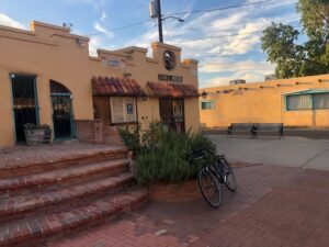 Exterior of a southwestern-style building with tan walls and red tile awnings. A bicycle is parked in front of brick steps leading to the entrance. The building appears to be a local business, with a sign visible above the door. The scene is set against a backdrop of a partly cloudy sky at sunup.