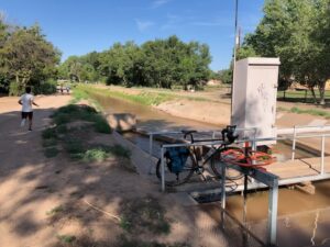 An irrigation ditch runs through a park-like area with trees. A person jogs on a path beside the water. In the foreground, a small metal bridge crosses the canal, with a bicycle parked on it. A white utility box stands next to the bridge.