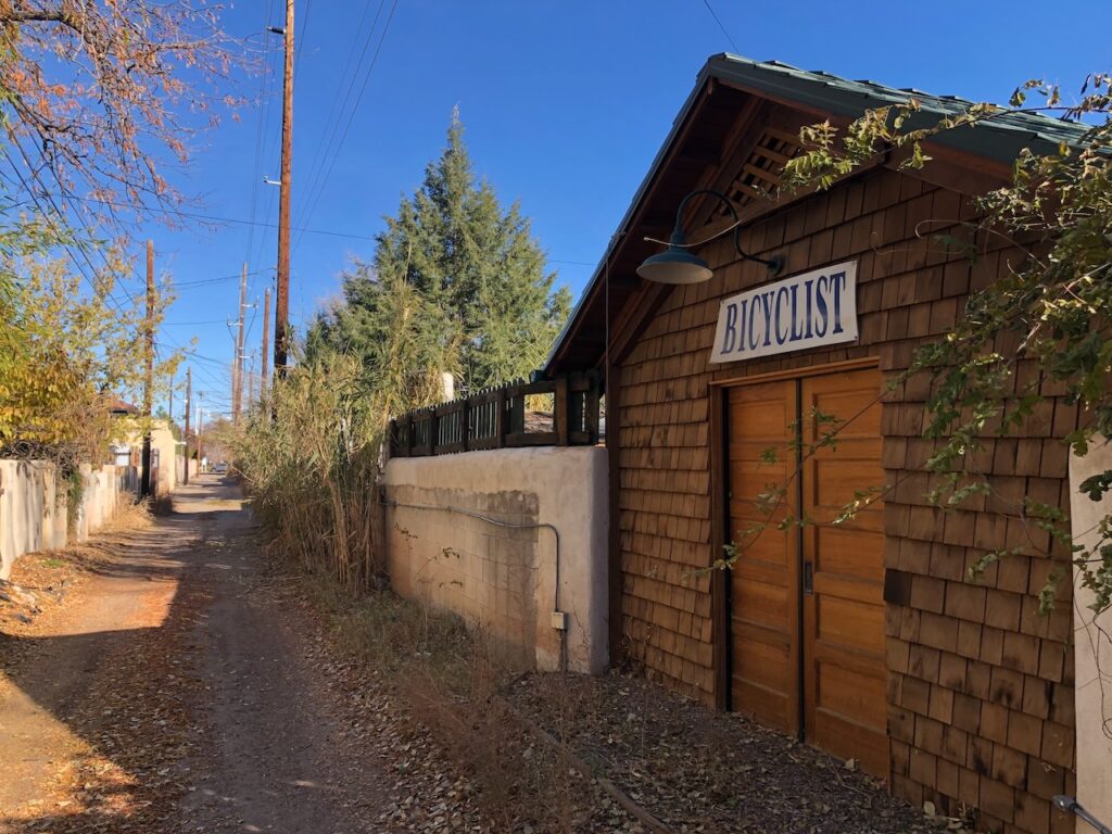 A dirt alley with fallen autumn leaves, featuring a cedar-shingled building with a sign reading "BICYCLIST" mounted on its wall. The building has wooden double doors and a gooseneck lamp above. Utility poles and bamboo line the alley, and the scene is lit by warm sunlight under a bright blue sky.