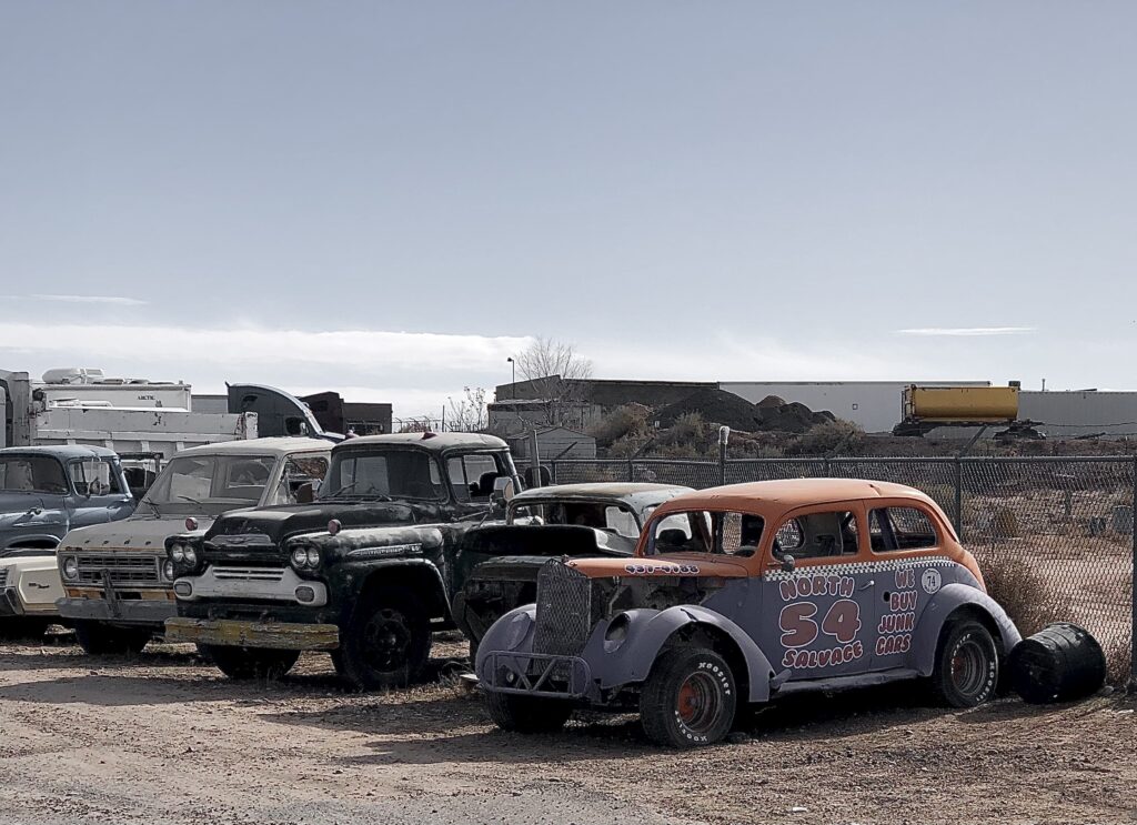 "An outdoor lot filled with old, worn-out vehicles including vintage trucks, a painted salvage race car, and other aged automobiles. The salvage race car is painted in bright orange and purple, with 'North 54 Salvage' and 'We Buy Junk Cars' on the side. The background includes an industrial yard with construction equipment and a dirt mound."