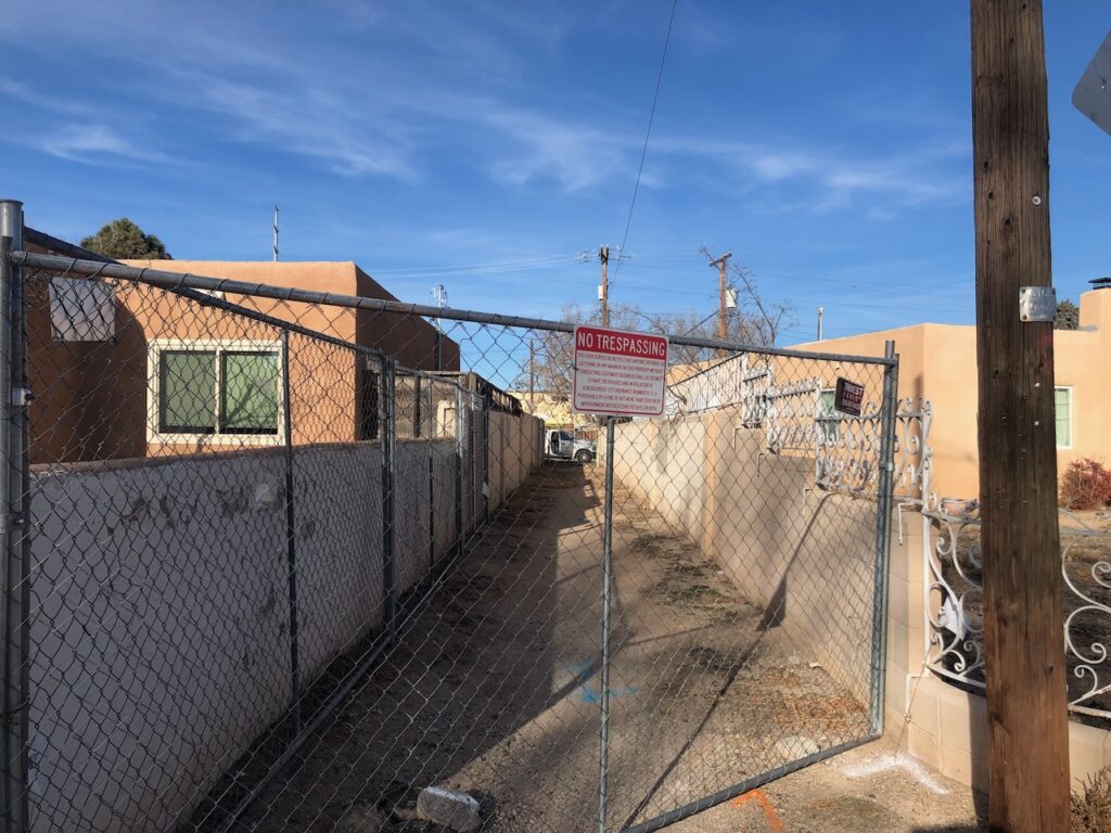 Temporary fence across a narrow dirt alley, with cinder block walls and houses on either side.