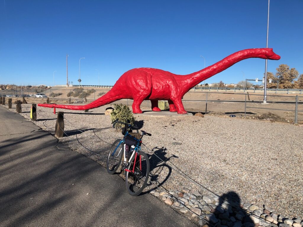 Large red dinosaur sculpture surrounded by a low fence with a bicycle leaning against the fence and the shadow of the photographer poking into the bottom right corner of the picture.