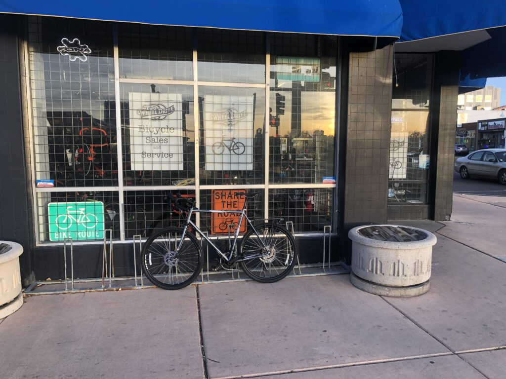 A silver-grey bicycle sitting on the sidewalk in front of a bike shop.