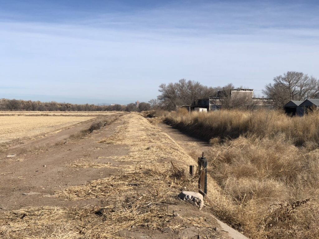 Brown farm field bisected by ditch and service road with old brick building on the right.