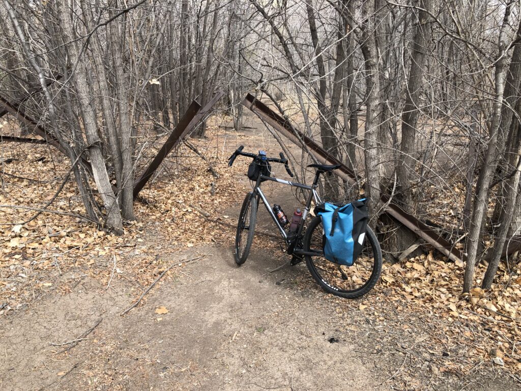 Bicycle leaning against rusted steel structure that looks like a giant version of a child’s jacks, with dirt trail and trees.