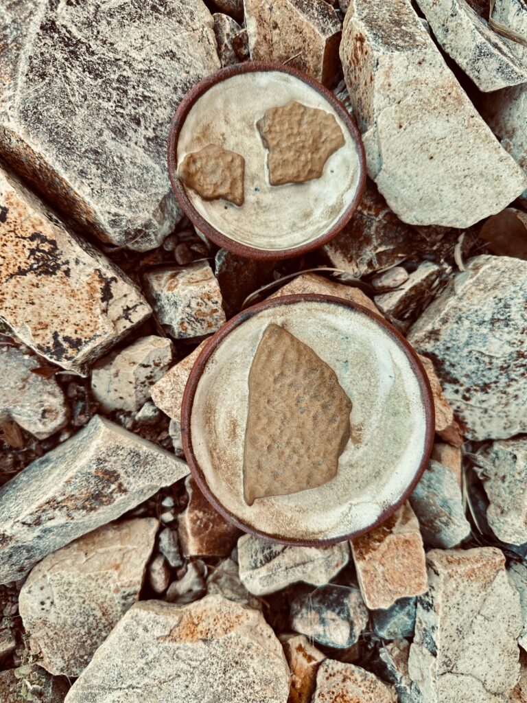 Two small ceramic plates, set on a background of rocks. Each plate includes fired pieces of cracked mud.