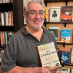 Gray haired, gray bearded man standing before a bookstore shelf holding a book entitled "The Crossing."