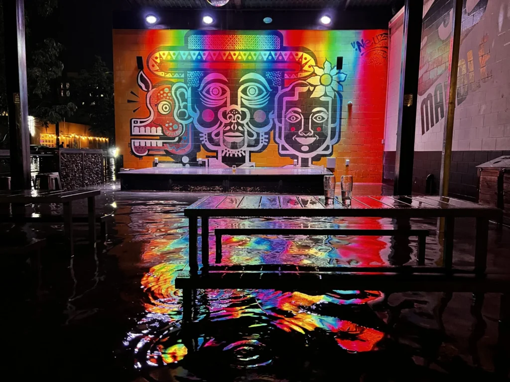 A vibrant nighttime scene featuring a colorful mural on a wall. The mural depicts three stylized faces with abstract designs in rainbow colors. In the foreground, a wet surface reflects the mural's colors, creating a kaleidoscopic effect. Spotlights illuminate the mural, and a table is visible in the foreground.
