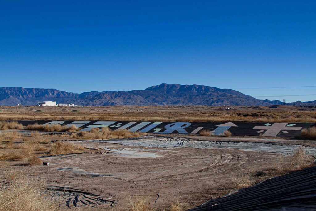 A vibrant desert scene under a flawless blue sky, featuring graffiti-like markings on a large black tarp sprawled across the dry, scrubby landscape. In the distance, the rugged Sandia Mountains rise dramatically, framed by a few scattered structures that hint at human presence amidst the raw beauty of the terrain.